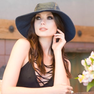 May include: A woman with long brown hair wearing a dark blue denim hat and a black sleeveless top with criss-cross straps.