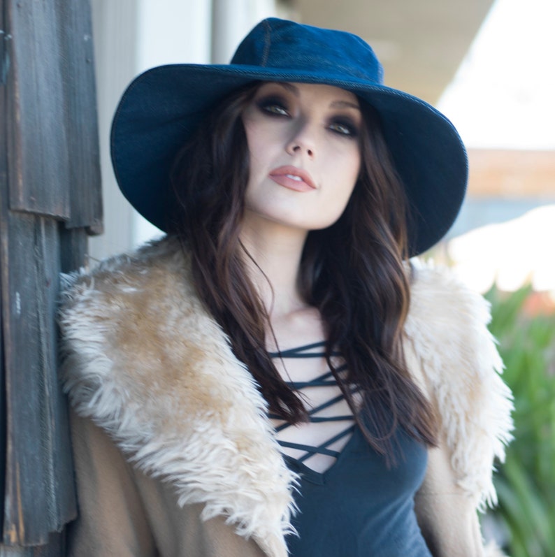 May include: A woman wearing a dark blue wide-brimmed hat and a tan coat with a cream-coloured faux fur collar. The woman is wearing a black top with a criss-cross neckline.