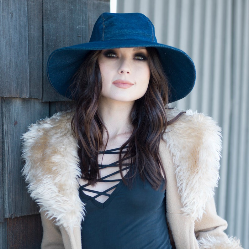 May include: A woman wearing a dark blue denim sun hat with a wide brim. She is also wearing a black top with a criss-cross neckline and a tan faux fur coat. The background is a wooden wall and a corrugated metal wall.
