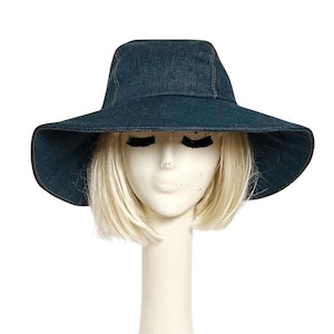 May include: A dark blue denim sun hat with a wide brim. The hat is displayed on a mannequin head with blonde hair. The hat has a classic shape, with a slightly rounded crown and a wide, floppy brim, perfect for sun protection.