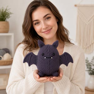 Könnte beinhalten: Ein dunkelviolettes gestricktes Fledermaus-Stofftier mit zwei spitzen Ohren, zwei ausgebreiteten Flügeln, kleinen Füßen, schwarzen Perlenaugen und zwei kleinen weißen Fangzähnen, die aus einem gestickten Lächeln hervorragen.