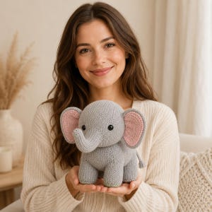 May include: A gray knitted elephant plush toy with large ears featuring light pink inner panels, a curved trunk, and two small black bead eyes. The stuffed animal has a textured yarn surface and a small tail with a tassel at the end.