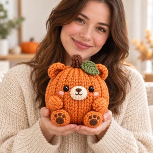 May include: A round, orange, knitted plush toy shaped like a pumpkin with bear features, including small ears, a cream-colored muzzle, black eyes, a brown stem, and a green leaf. The feet have dark brown paw pads, and the cheeks have pink accents.