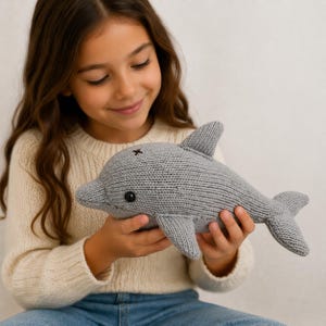 Könnte beinhalten: Ein graues, gestricktes Delfin-Plüschtier mit einem schwarzen Auge und einem kleinen schwarzen Kreuz auf dem Kopf. Der Delfin hat Flossen und einen Schwanz. Das Spielzeug wird von einer Person gehalten, die einen cremefarbenen Pullover und blaue Jeans trägt.