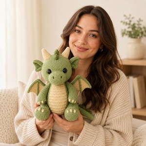May include: A knitted green dragon plush toy with a cream-colored belly, inner wings, and horns. The creature features black plastic eyes, a textured knit surface, and a small tail. The dragon is held in a seated position with visible claws.