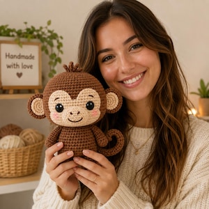 Könnte beinhalten: Ein gehäkeltes Affen-Plüschtier in Braun und Beige mit schwarzen Sicherheitsaugen, einem kleinen Haarbüschel auf dem Kopf und rosa gestickten Wangen. Der Affe hat einen geringelten Schwanz, runde Ohren und einen gestickten Mund aus Garn.