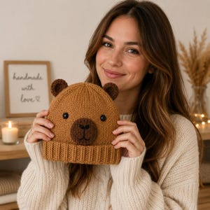May include: A brown knitted beanie featuring a bear face design with two small rounded ears on top, a dark brown circular snout, and two black button eyes. The hat has a ribbed cuff at the bottom and is constructed from a textured yarn material.