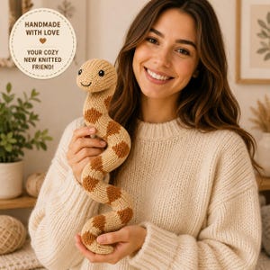 May include: A knitted snake toy featuring a light beige and brown patterned design with black embroidered eyes and a small smile. A circular label is visible with the text: HANDMADE WITH LOVE, YOUR COZY NEW KNITTED FRIEND!