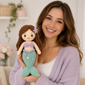 Puede incluir: Una muñeca sirena tejida con cabello de lana marrón, un top de concha morado y una cola verde menta. La muñeca presenta una pequeña flor morada en el cabello y detalles florales bordados en la cola. Tiene un rostro color crema con ojos negros.
