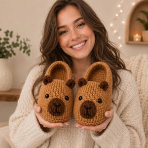 Puede incluir: Un par de pantuflas de ganchillo color canela con forma de capibaras, que presentan narices circulares de color marrón oscuro, orejas pequeñas y redondeadas y ojos de cuentas negras. Tienen un patrón de tejido texturizado y cerrado.