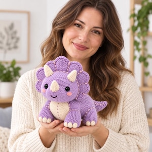Puede incluir: Un juguete de dinosaurio triceratops tejido en color morado claro con vientre, cuernos y pies de color crema. El juguete presenta un gran volante alrededor de la cabeza, ojos de botón negros y pequeños círculos rosas bordados.