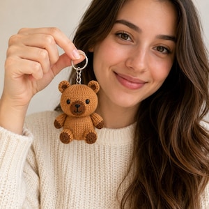 May include: A small, brown, knitted teddy bear keyring featuring a round head, two small ears, and dark brown accents on the paws and snout. The bear has two black bead eyes and a stitched nose and mouth, attached to a silver-toned metal keyring.