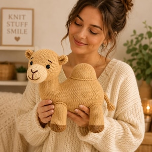 May include: A tan knitted camel plush toy with a lighter beige muzzle, dark brown embroidered facial features, and dark brown feet. The camel has a single hump on its back and a small tail, crafted with a visible knit texture throughout the body.