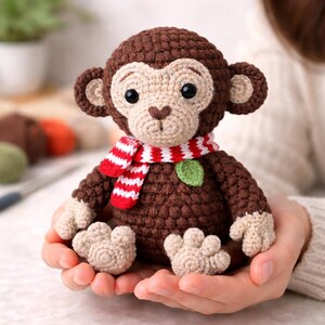 May include: A handmade crochet monkey doll with a brown body, tan face, and a red and white striped scarf. The monkey has black eyes and is holding a green leaf. The doll is a cute, soft toy.