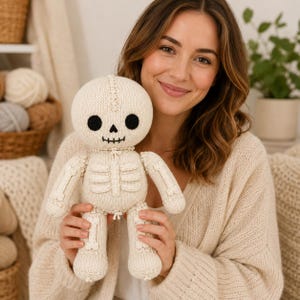 May include: A cream-coloured knitted skeleton doll featuring a round head with black circular eyes, a small triangular nose, and a stitched mouth. The body displays a rib cage and limb bone details crafted from matching yarn, with a central seam.