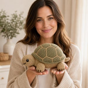 May include: A knitted turtle toy featuring a tan head, legs, and tail with a textured olive green shell. The shell has a distinct beige stitched hexagonal pattern. The turtle has small black embroidered eyes and a simple curved black mouth line.