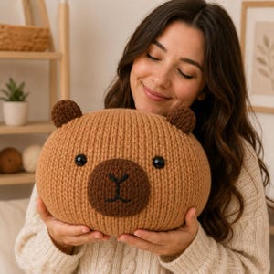 Peut inclure: Une peluche tricotée ronde de couleur brun clair en forme de capybara. Elle présente un museau circulaire brun foncé avec un nez et une bouche brodés en noir, deux petites oreilles brun foncé et deux yeux ronds en plastique noir sur la texture côtelée.