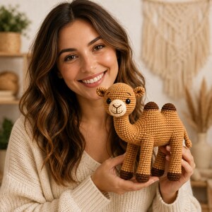 May include: A tan crochet camel stuffed animal featuring two humps, dark brown hooves, and a light cream-colored muzzle. The toy has black safety eyes and a stitched mouth, crafted with a textured yarn finish in a standing position.