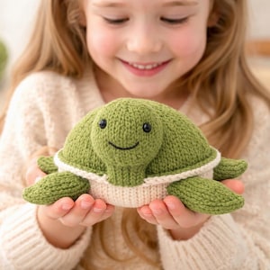 Puede incluir: Un peluche de tortuga verde tejido con una cara sonriente, ojos negros y ribete blanco. La tortuga está sostenida en las manos de una persona. Es un juguete blando.