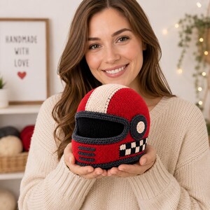 May include: A crocheted motorcycle helmet toy in bright red with a cream-colored center stripe, a black visor area, and a black and white checkered pattern on the side. The helmet features dark gray trim and three horizontal dark gray lines.