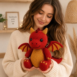 May include: A knitted dragon plush toy featuring a vibrant red body with contrasting mustard yellow accents on the belly, inner wings, and feet. The creature has two black plastic eyes, a small stitched smile, and a spiky ridge along its back.