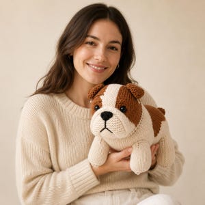 May include: A handmade knitted bulldog plush toy featuring a cream-coloured body with brown patches on the head and back, black plastic eyes, and a black stitched nose. The stuffed animal has a textured yarn finish and a sturdy, standing posture.