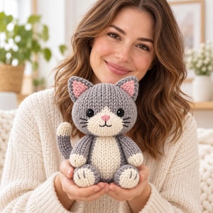 Op de afbeelding: Een klein, handgebreid pluche kattenspeeltje met een grijs lichaam en een crèmekleurige buik, snuit en voetzooltjes. De kat heeft roze binnenoren, een roze neus, zwarte geborduurde ogen en snorharen, met een gekrulde staart aan de linkerzijde.