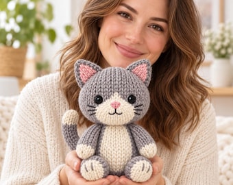 May include: A small, hand-knitted plush cat toy featuring a grey body with a cream-colored belly, muzzle, and paw pads. The cat has pink inner ears, a pink nose, black embroidered eyes, and whiskers, with a curved tail extending to the left side.