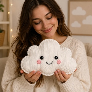 May include: A cream-colored, cloud-shaped plush pillow featuring a knitted texture, two black circular eyes, a stitched black smile, and two small, round, pink textured cheeks. The edges of the cloud are finished with a thick, visible blanket stitch.