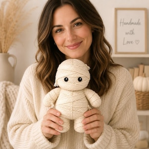 Puede incluir: Una pequeña muñeca momia tejida a mano en color crema, con un diseño de vendas envueltas, ojos negros redondos y una textura de hilo suave. La muñeca tiene una cabeza redondeada, brazos y piernas cortos con extremos de hilo visibles.