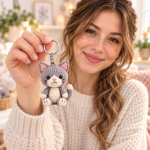 Peut inclure: Un porte-clés chat crocheté gris et crème avec un petit anneau en métal. Le chat a des oreilles intérieures roses, des yeux brodés noirs, un nez rose et une bouche cousue. Le corps est gris avec un ventre, des pattes et une queue gris.