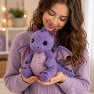 May include: A knitted purple dragon plush toy featuring a lighter purple belly, dark purple inner wings, and dark purple horns. The creature has black safety eyes, a textured knit surface, and a long tail with small spikes along the back.