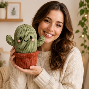 Puede incluir: Un cactus de peluche tejido con un cuerpo verde salvia con dos brazos laterales, una maceta de color naranja oscuro y una cara sonriente con ojos de cuentas negras y pequeñas mejillas rosadas bordadas. La textura es de punto en todo el objeto.