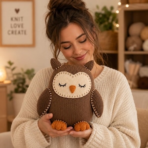 Op de afbeelding: Een bruine gebreide uil knuffel met een crèmekleurig gezicht, gesloten ogen met zwarte geborduurde wimpers en een oranje snavel. De uil heeft oranje voeten en decoratieve crèmekleurige stiksels langs de vleugels en de rand van het gezicht.