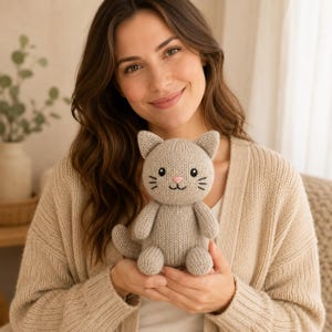 Può includere: Un piccolo peluche a forma di gatto lavorato a maglia color beige chiaro con testa arrotondata, orecchie a punta e un semplice viso ricamato con occhi neri, naso rosa e bocca cucita. Il peluche ha una superficie in filato morbido e testurizzato.