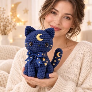 May include: A dark blue crocheted cat plush toy featuring a yellow crescent moon on its forehead, large black eyes, and a dark blue satin ribbon tied around its neck. The cat is decorated with small yellow embroidered stars and a moon on its tail.