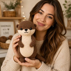 May include: A brown and cream-colored crochet otter plush toy with a rounded head, small ears, black plastic eyes, and a stitched nose and mouth. The stuffed animal features a textured yarn construction with a lighter cream belly and dark brown limbs.