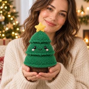 May include: A small, hand-knitted green Christmas tree plush toy with a brown base and a yellow star on top. The tree features a friendly embroidered face with black bead eyes, a small smile, and pink cheeks, crafted from soft yarn.