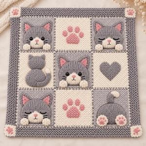 May include: A square knitted blanket featuring a grid of nine panels in gray and cream. The design includes raised 3D motifs of gray cats with pink ears, pink paw prints, and a gray heart. The border is gray with small pink paw prints at corners.