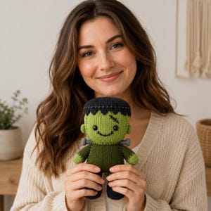 May include: A small, handmade knitted doll resembling Frankenstein's monster. The doll features a bright green head with black hair, a stitched scar, black button eyes, and a smiling mouth. It has a dark green body, grey neck bolts, and black feet.