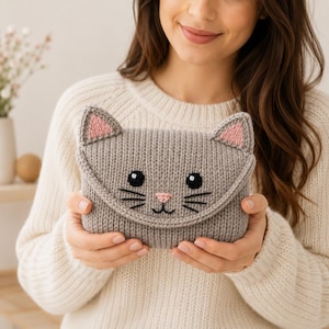 May include: A grey knitted pouch shaped like a cat face with a triangular flap. The pouch features two pointed ears with pink centers, two black embroidered eyes, a small pink embroidered nose, and black whiskers stitched onto the front.