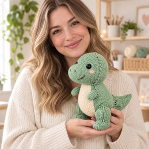 Può includere: Un dinosauro di peluche lavorato a maglia con corpo verde salvia, pancia color crema, braccia piccole, coda spessa, occhi a bottone neri e piccole guance ricamate in rosa. Il giocattolo ha una testa arrotondata e una trama a maglia visibile.