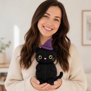 May include: A black crocheted cat plush toy featuring a small purple witch hat, bright yellow eyes with black pupils, and a tiny pink nose. The cat has a rounded body, small ears, and a curved tail, all crafted with a visible knit texture.