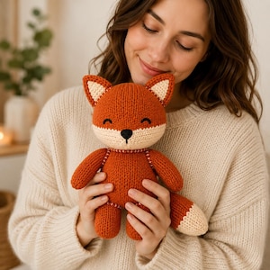 Puede incluir: Un peluche de zorro tejido con un cuerpo de color naranja vibrante, con el hocico, el interior de las orejas y la punta de la cola en color crema. El zorro tiene una pequeña nariz negra bordada y ojos cerrados con líneas curvas.