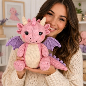 May include: A knitted plush dragon toy featuring a soft pink body, a cream-colored belly, and purple wings. The dragon has two small cream horns, black button eyes with long eyelashes, and a row of small purple spikes running down its long tail.