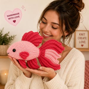 May include: A crocheted fish toy featuring a light pink body with dark pink fins and a large, flowing dark pink tail. The fish has a small black eye and a stitched mouth. The text "HANDMADE WITH LOVE" is visible in a heart-shaped graphic above the fish.