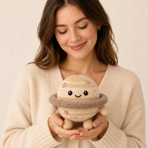 May include: A round, beige, knitted planet plush toy featuring a darker brown ring around its center, small arms, and two legs. The plush has a smiling face with two black embroidered eyes and a small curved mouth, along with light brown spots.
