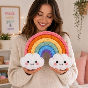 Pode incluir: Uma almofada de arco-íris em crochê com um arco multicolorido em rosa, laranja, amarelo, azul-petróleo e roxo. O arco-íris é ancorado por duas nuvens brancas de pelúcia sorridentes com olhos pretos e bochechas rosadas, em fio texturizado.