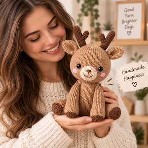 May include: A light brown knitted deer plush toy with dark brown antlers, black eyes, a white muzzle, and pink cheeks. The toy features a small embroidered nose and mouth. A tag attached to the side displays the text Handmade Happiness with a heart.