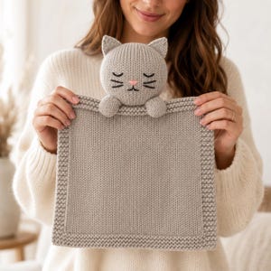 Puede incluir: Una manta de seguridad tejida en color gris claro con una cabeza de gato que presenta ojos cerrados, bigotes y una nariz rosa bordados. La manta cuadrada tiene un borde texturizado y dos pequeñas patas de gato en el borde superior.